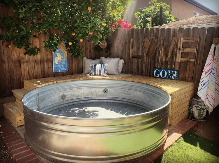 A large galvanized steel tub filled with water, placed outdoors in a backyard setting with wooden steps, cushions, a fence, and garden plants by stock tank pools wa